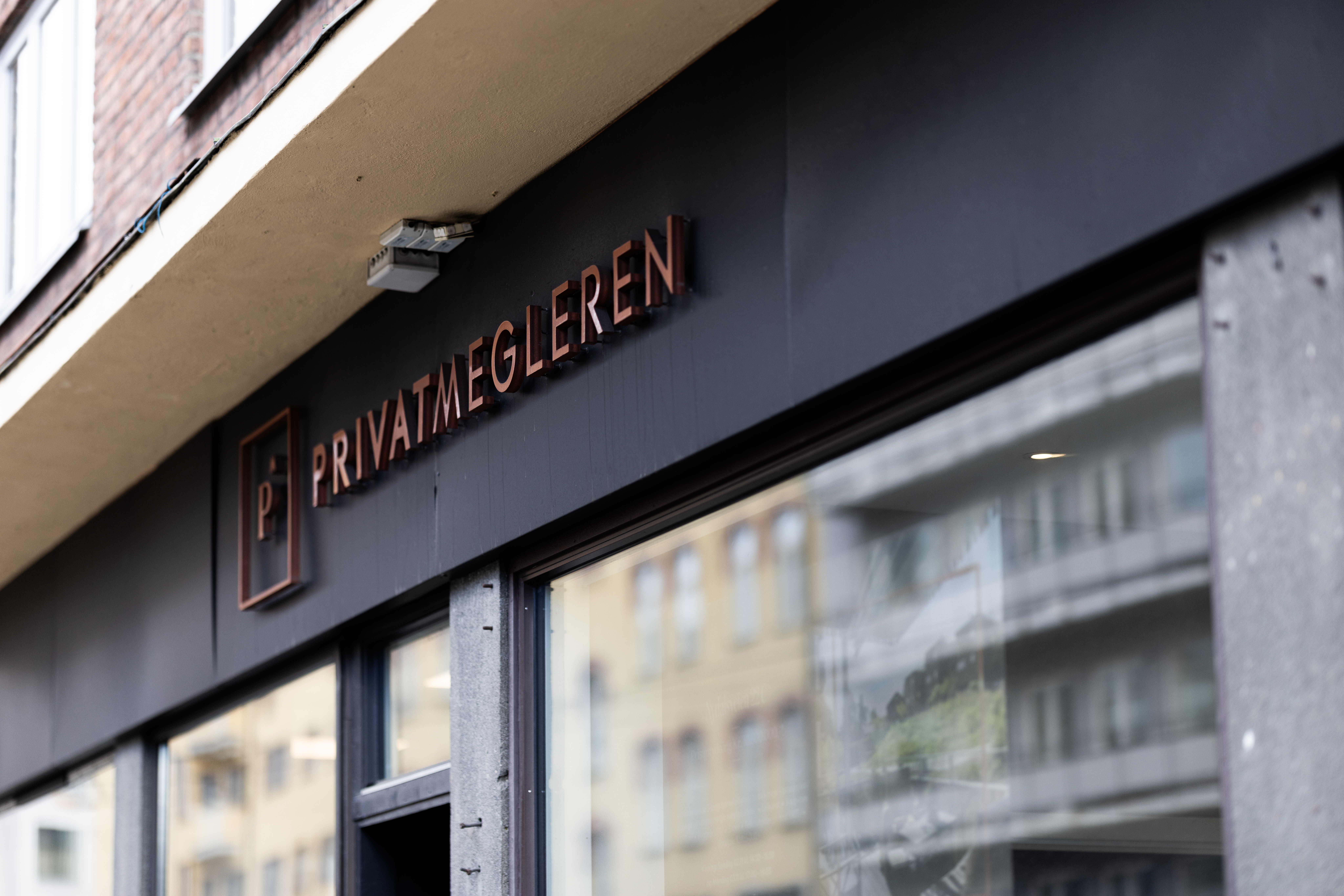 PrivatMegleren-skilt på mørk fasade med vindusspeiling av bygårder, brukt som illustrasjon i artikkel om eiendomsmegler på Vollebekk.